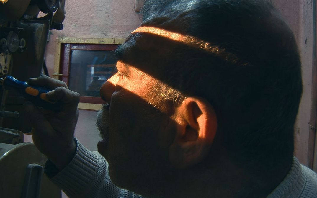 Film still from HABIBI HUSSEIN: An older man is tinkering with an analog film projector. The room is dark, with sunlight streaming in through a gap near his eyes.