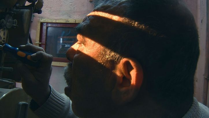 Film still from HABIBI HUSSEIN: An older man is tinkering with an analog film projector. The room is dark, with sunlight streaming in through a gap near his eyes.