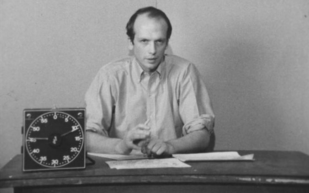 Filmstill aus PICTURE AND SOUND RUSHES: Ein Mann in einem Hemd sitzt an einem Tisch und erklärt etwas. Vor ihm liegen Blätter und eine Uhr steht auf dem Tisch.