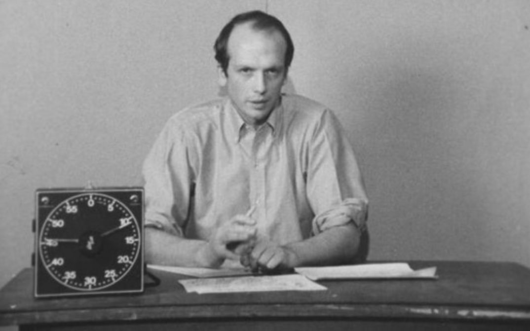 Film still from PICTURE AND SOUND RUSHES: A man in a shirt is sitting at a table, explaining something. There are some sheets of paper in front of him, and a clock is on the table.