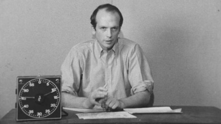 Filmstill aus PICTURE AND SOUND RUSHES: Ein Mann in einem Hemd sitzt an einem Tisch und erklärt etwas. Vor ihm liegen Blätter und eine Uhr steht auf dem Tisch.