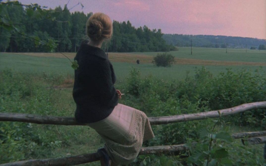 Film still from ZERKALO: A woman sits on a fence and looks into the wide, green landscape. 
