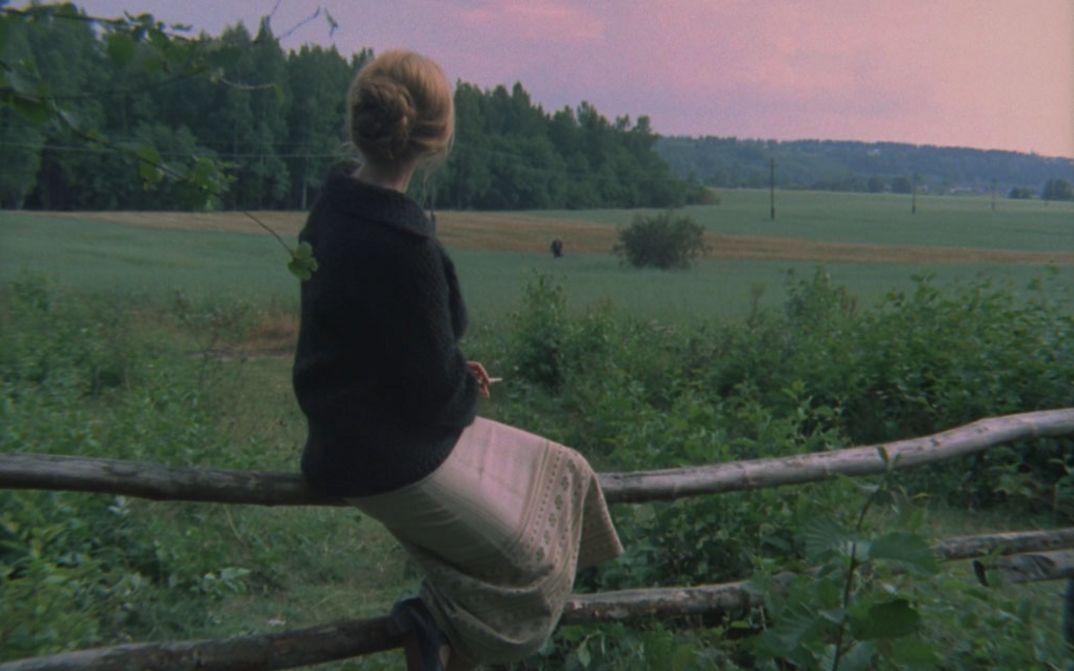 Film still from ZERKALO: A woman sits on a fence and looks into the wide, green landscape.