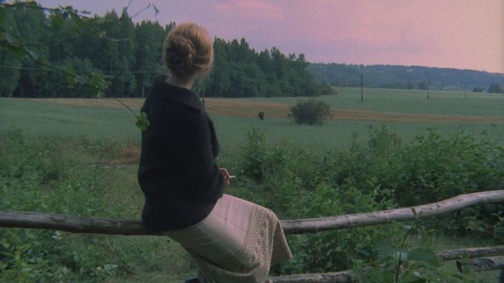 Film still from ZERKALO: A woman sits on a fence and looks into the wide, green landscape. 