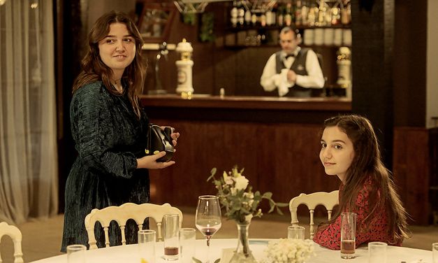 Filmstill from "Cidade Rabat" by Susana Nobre. A young woman is sitting at a table on which one guesses the end of a fancy meal. Another one is standing next to it. In the background is a bar counter. 