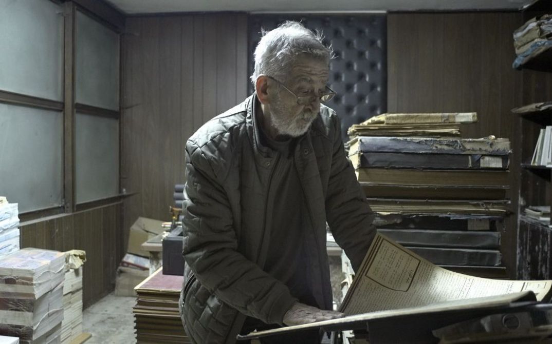 Ein älterer Mann steht zwischen Aktenstapeln und liest ein großes Dokument.