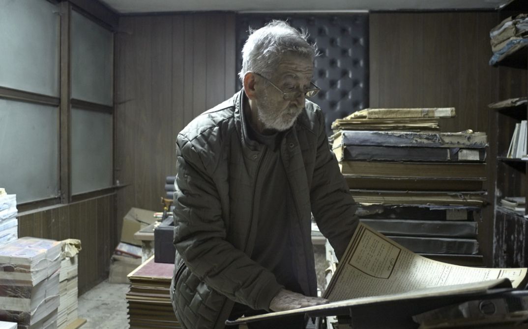 An elderly man stands among stacks of files, reading a large document.