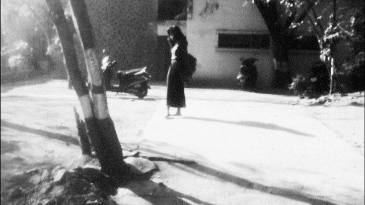 Filmstill aus A NIGHT OF KNOWING NOTHING: Man sieht in schwarz-weiß eine leicht verschwommene Straßenszene. Im Vordergrund stehen zwei Bäume. Auf der Straße steht eine Frau mit langen schwarzen Haaren. Die Sonne wirft ein helles Licht und harte Schatten.