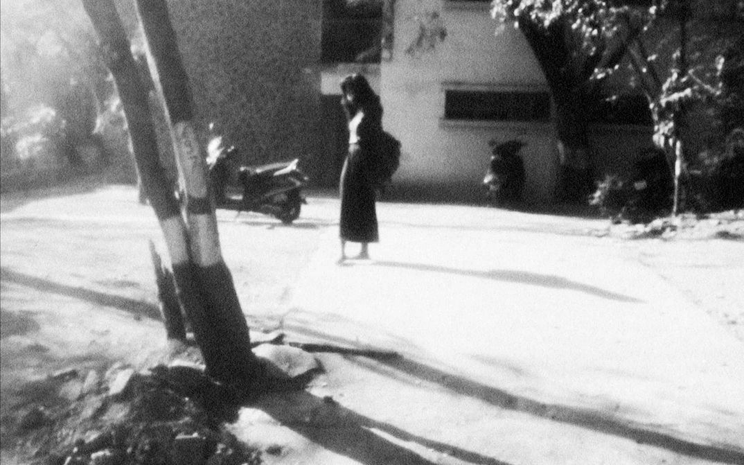 Filmstill aus A NIGHT OF KNOWING NOTHING: Man sieht in schwarz-weiß eine leicht verschwommene Straßenszene. Im Vordergrund stehen zwei Bäume. Auf der Straße steht eine Frau mit langen schwarzen Haaren. Die Sonne wirft ein helles Licht und harte Schatten.