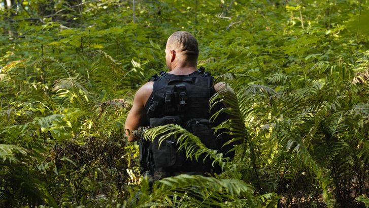Film still from LES LIGNES ENEMIES: A muscular man in combat gear can be seen from behind. He is standing in a forest surrounded by ferns.