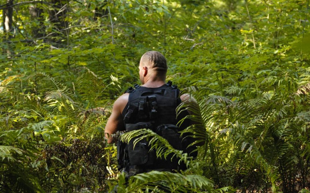 Filmstill aus LES LIGNES ENEMIES: Man sieht von hinten einen muskulösen Mann in Kampfmontur. Er steht in einem Wald inmitten von Farnen.