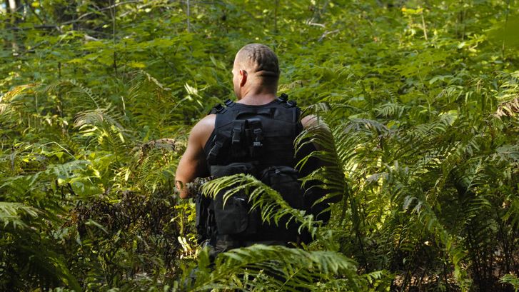 Filmstill aus LES LIGNES ENEMIES: Man sieht von hinten einen muskulösen Mann in Kampfmontur. Er steht in einem Wald inmitten von Farnen.