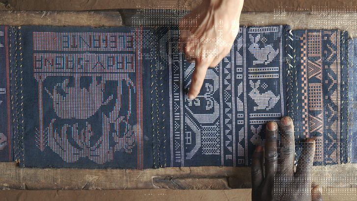 Film still from QUANTUM CREOLE: View of an elaborately woven cloth. A black and a white hand point to it.