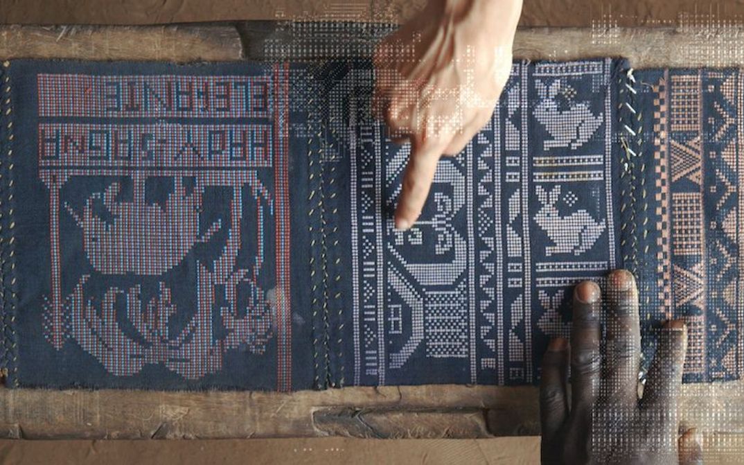 Filmstill aus QUANTUM CREOLE: Ansicht eines aufwendig gewebten Tuchs. Eine schwarzen und eine weiße Hand zeigen darauf.