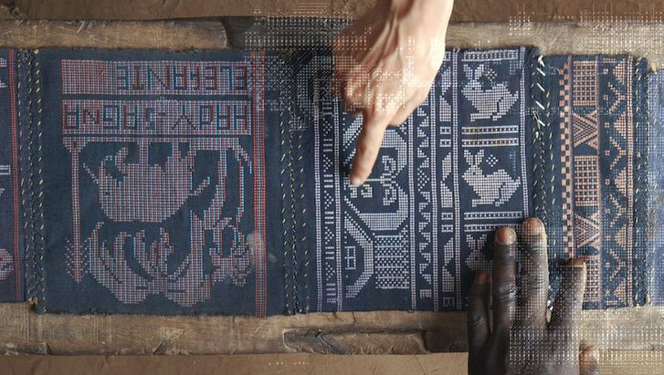 Filmstill aus QUANTUM CREOLE: Ansicht eines aufwendig gewebten Tuchs. Eine schwarzen und eine weiße Hand zeigen darauf.