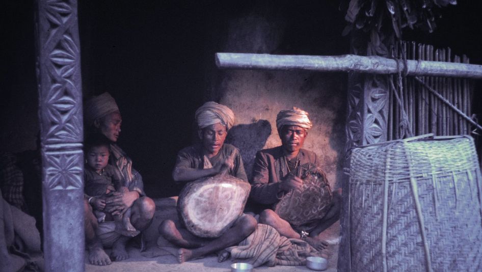 Film still from Shamans of the Blind Country: Three men are sitting next to each other on the floor. Two are drumming on small drums, one has a small child on his lap.