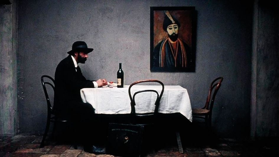 Filmstill aus PIROSMANI: Ein schwarz angezogener Mann mit Hut sitzt alleine an einem Esstisch. An der Wand hängt ein von Pirosmani gemaltes Bild.