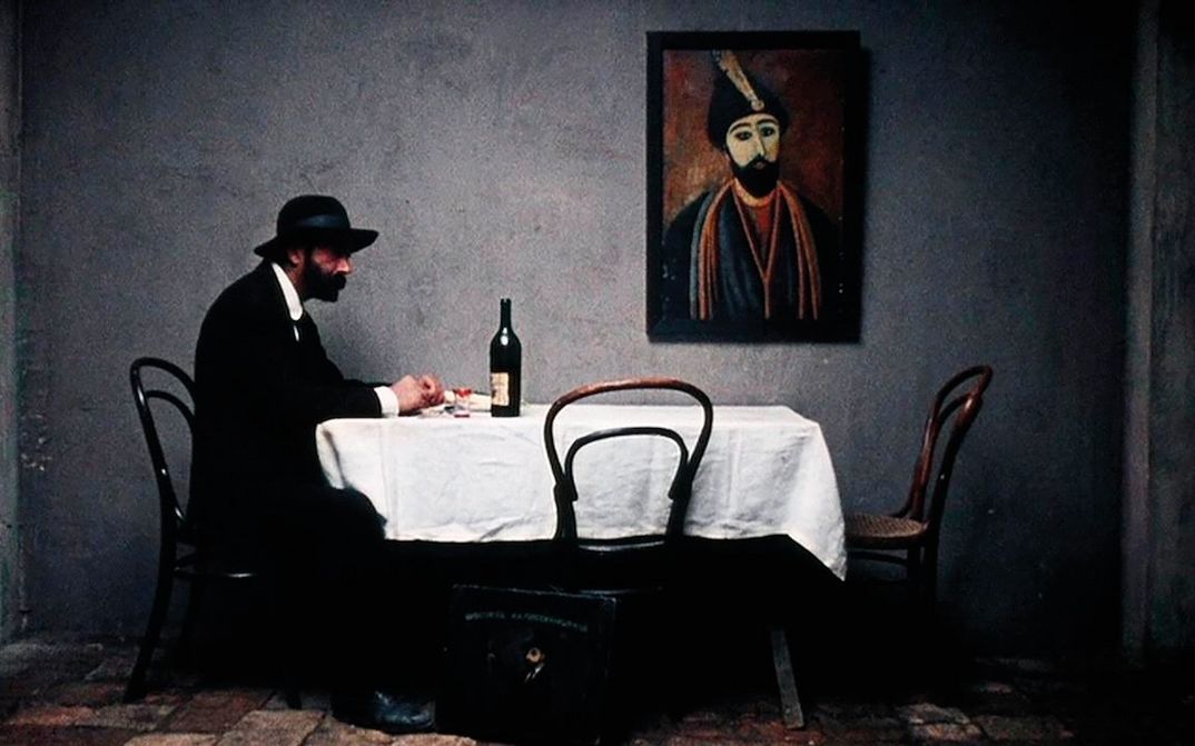 Film still from PIROSMANI: A man dressed in black and wearing a hat is sitting alone at a dining table. A painting by Pirosmani hangs on the wall.
