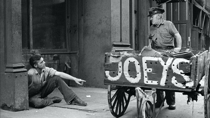 Filmstill aus ON THE BOWERY: Ein Mann zieht einen großen Handkarren, auf dem "Joeys" geschrieben ist, durch die Straße. Ein anderer Mann sitzt auf dem Gehweg. Beide blicken sich an und reden miteinander.