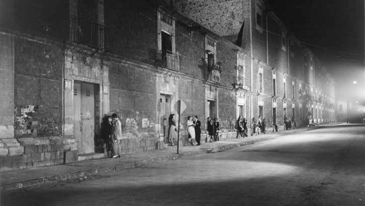 Film still from TROTACALLES: Scattered sex workers and couples stand along a sparsely lit street.