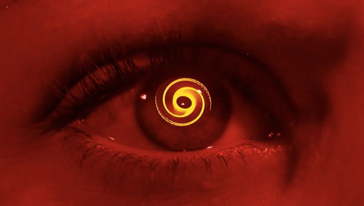 Filmstill aus VERTIGO: Monochrom rot eingefärbte Nahaufnahme eines Auges, in der Pupille dreht sich eine Spirale.