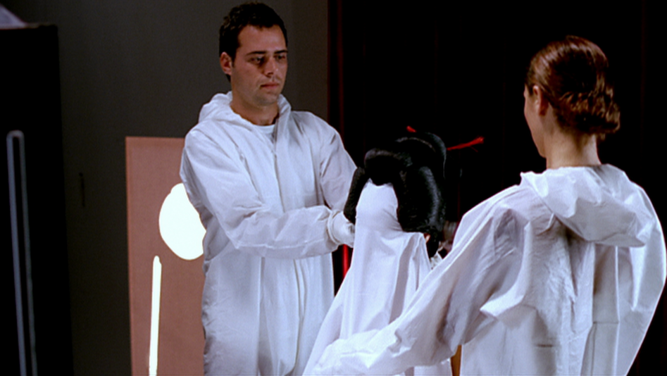 Film still from ANAESTHETICS: Two people in white hooded overalls are working on a figure sitting between them.