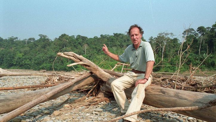 Film still from MEIN LIEBSTER FEIND: Werner Herzog sits on a lying tree trunk and points to the jungle behind him. 