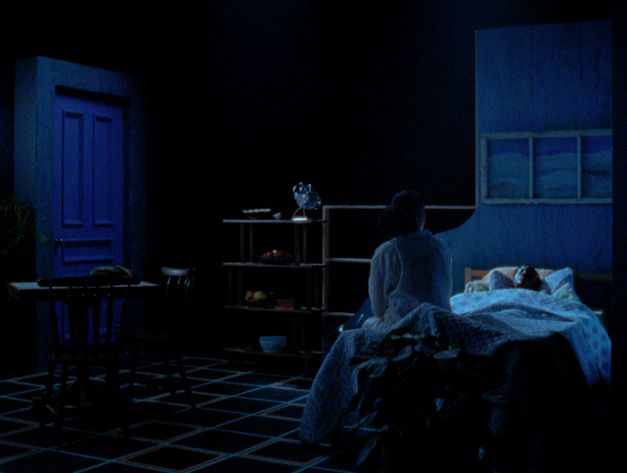 Still from the film "Cette Maison (This House)" by Miryam Charles. A girl lays in bed in a bedroom on a dark soundstage, with a woman sitting at the foot of the bed.