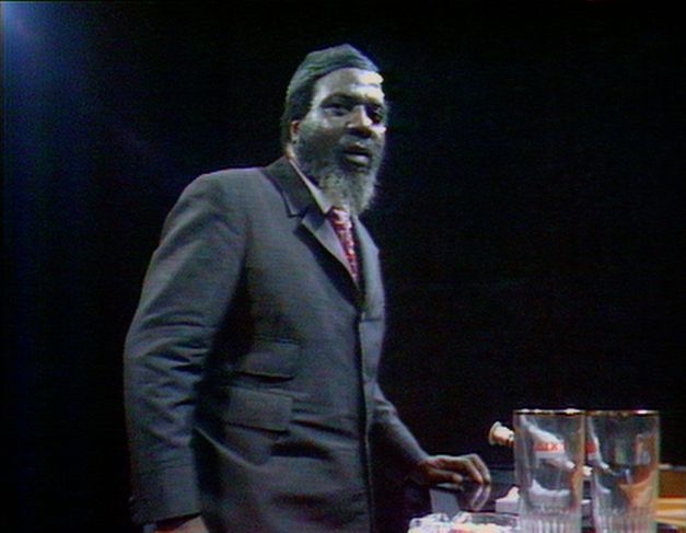 Filmstill aus „Rewind & Play“. Eine Video-Aufnahme. Thelonious Monk steht vor dunklem Hintergrund vor seinem Klavier. Ein paar Wassergläser stehen auf dem Klavier.