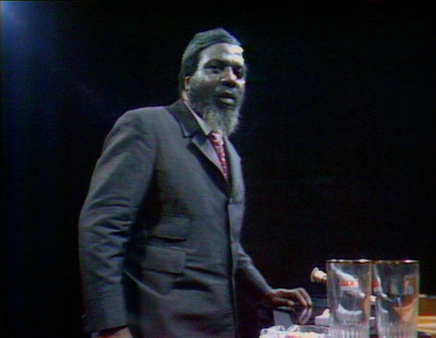 Still from the film “Rewind & Play“. A video image of Thelonious Monk standing up in front of a piano. The background is dark, there are some glasses of water on the piano in the foreground.