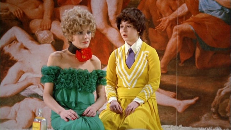 Filmstill aus DIE BITTEREN TRÄNEN DER PETRA VON KANT: Eine Frau und ein Mädchen knien auf einem plüschigen Teppich. Die Frau trägt ein grünes Abendkleid und blondes lockiges Haar. Das Mädchen trägt einen gelben Rock und einen gelben Pullover. Neben der Frau steht eine Flasche Alkohol auf dem Boden. Die Wand im Hintergrund ist mit einem Barockgemälde bedeckt.
