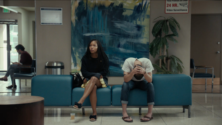 Film still from TEST PATTERN: A young couple sits side by side in a waiting room on a blue sofa.