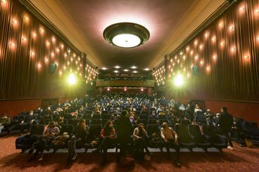 A full cinema, the photo is taken from the screen. Between the visitors, there is always a seat empty. Everyone is wearing masks.