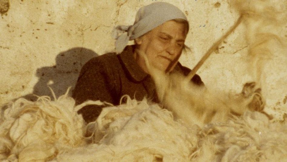 Filmstill aus Das fruchtbare Gedächtnis: Eine ältere Frau in Arbeitskleidung sitzt an einer Hauswand und bearbeitet vor ihr liegende Wolle.