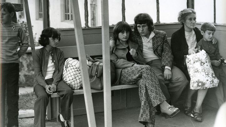 Filmstill aus EINFACHE GESCHICHTE: Einige Menschen sitzen auf einer Bank in einer Wartehalle. Im Zentrum ein junges Paar, sie schaut direkt in die Kamera.