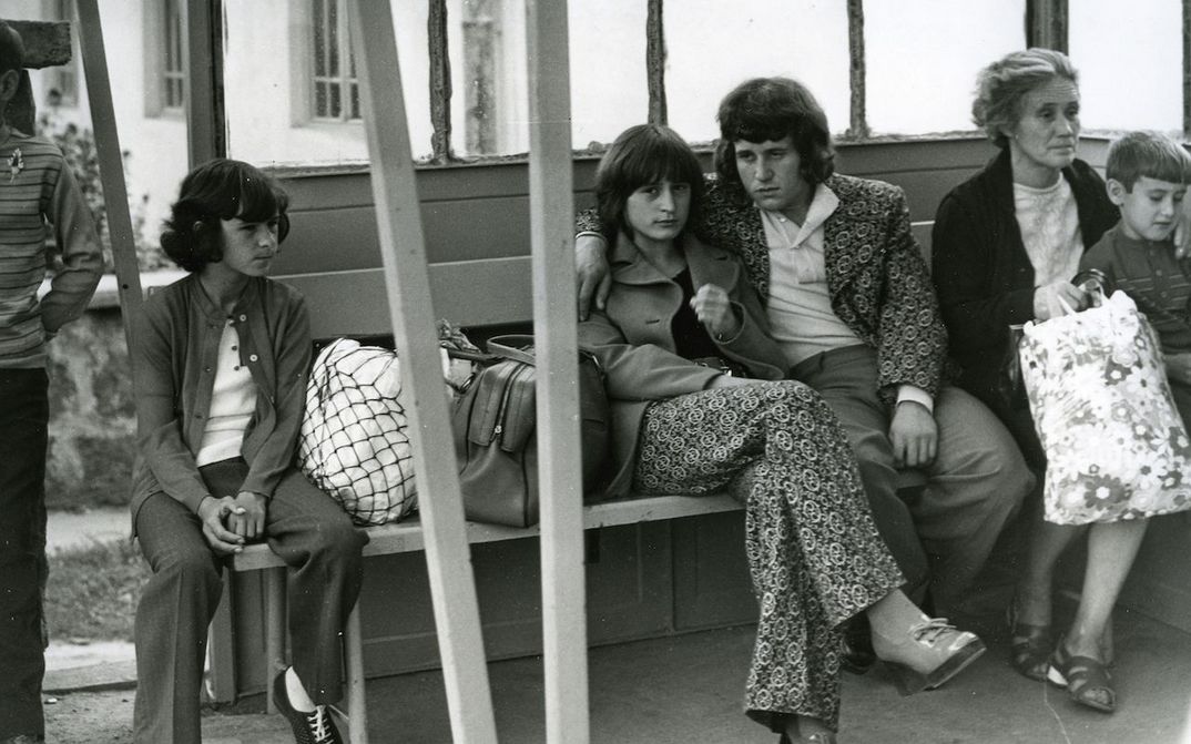 Filmstill aus EINFACHE GESCHICHTE: Einige Menschen sitzen auf einer Bank in einer Wartehalle. Im Zentrum ein junges Paar, sie schaut direkt in die Kamera.