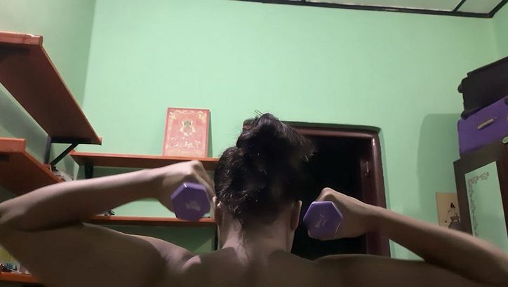 Still from the film "If from Every Tongue It Drips" by Sharlene Bamboat. A woman lifts two dumbbells in front of a green wall.