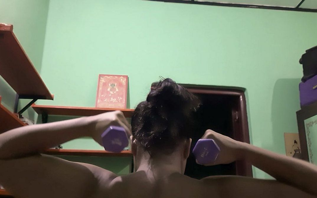 Still from the film "If from Every Tongue It Drips" by Sharlene Bamboat. A woman lifts two dumbbells in front of a green wall.