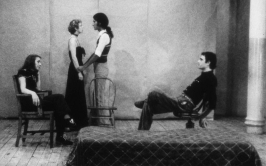 Film still from LIVES OF PERFORMERS: Four people standing and sitting on a stage.