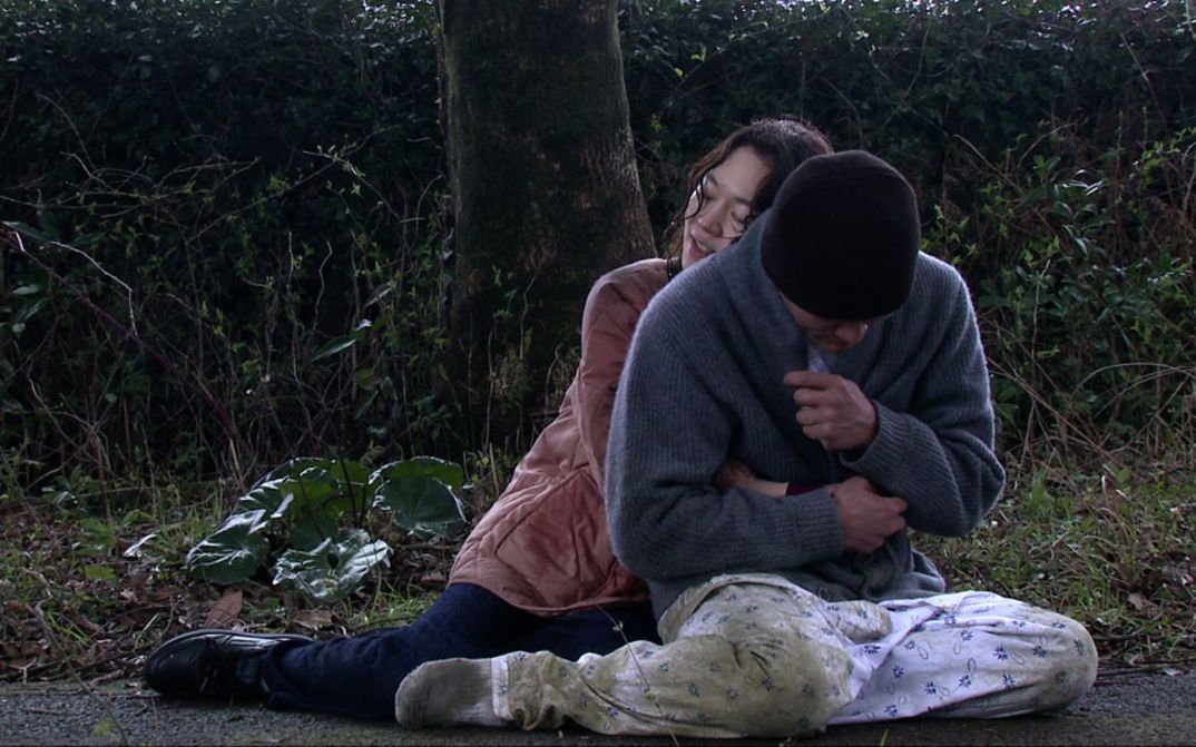 A man and a woman are sitting on the ground on a park path. She sits behind him and hugs him.