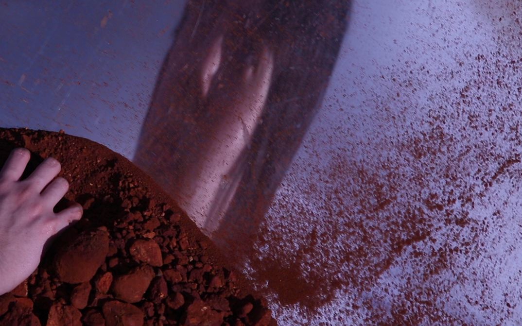 Film still from Sanna Gabraz’s film “Cartas do Absurdo”. A face is reflected on a mirrored surface, scattered with red earth that a hand in the foreground grasps.