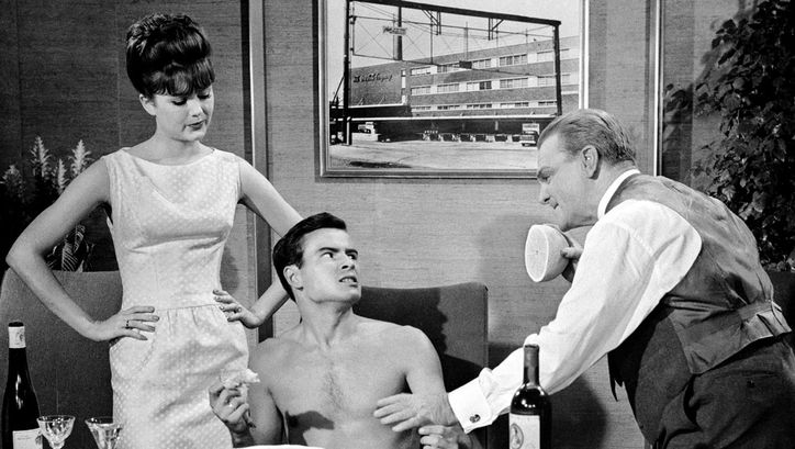 Film still from ONE, TWO, THREE: A young man with a naked upper body is sitting at a table eating. To his left and right are a woman and a man who are looking at him disapprovingly.