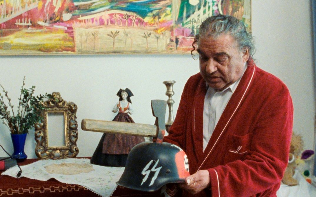 Filmstill aus WANKOSTÄTTN: Ein Mann mit grauem Haar und rotem Bademantel hält einen Helm mit SS-Schriftzug in der Hand. In dem Helm steckt eine Axt und rote Farbe ist drumherum.