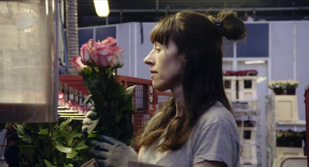 Still from the film "Une fleur à la bouche" by Éric Baudelaire. A woman wearing gloves in a warehouse holds a handful of white and pink roses.