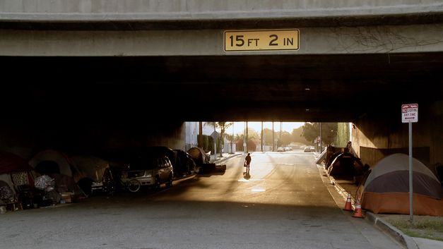 Still aus James Bennings „The United States of America“: Wir schauen durch eine Straßenunterführung. Auf der anderen Seite scheint die Nachmittagssonne. Unter der Brücke am Straßenrand Zelte, in denen Menschen leben. 