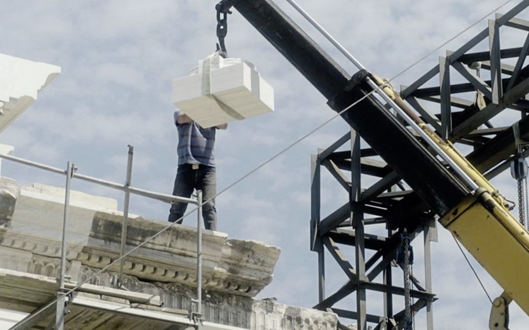 Filmstill aus „Onun Haricinde, yiyim“ von Eren Aksu. Eine Baustelle für ein Gebäude mit einem Kran. Ein Mann auf der Baustelle nimmt Bauelemente vom Kran entgegen.