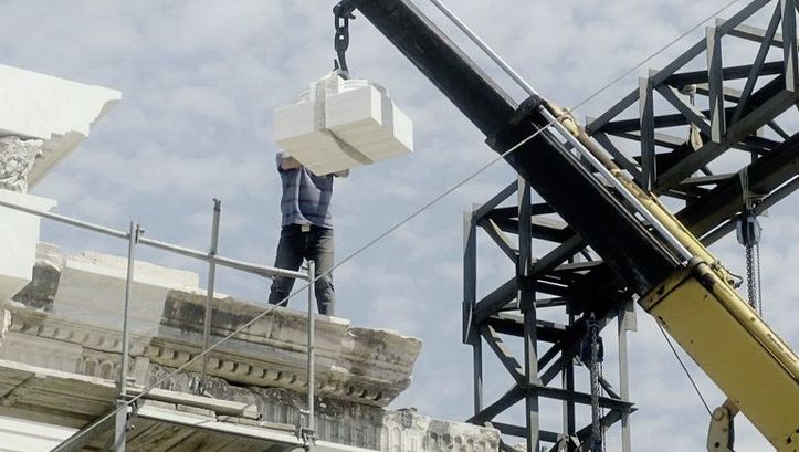 Still from the film „Onun Haricinde, yiyim“ by Eren Aksu. A construction site for a building with a crane. A man on the construction site receives building elements with the crane
