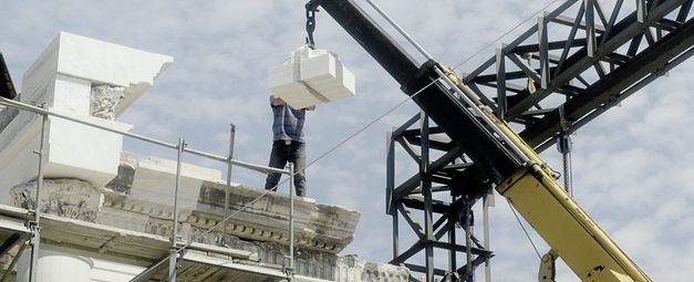 Still from the film „Onun Haricinde, yiyim“ by Eren Aksu. A construction site for a building with a crane. A man on the construction site receives building elements with the crane