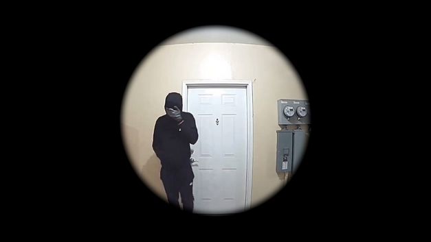 Film still from Graeme Arnfield’s „Home Invasion“. A circular view into a room. A door in the center; next to it a person dressed in black, obscuring their face with their hand.