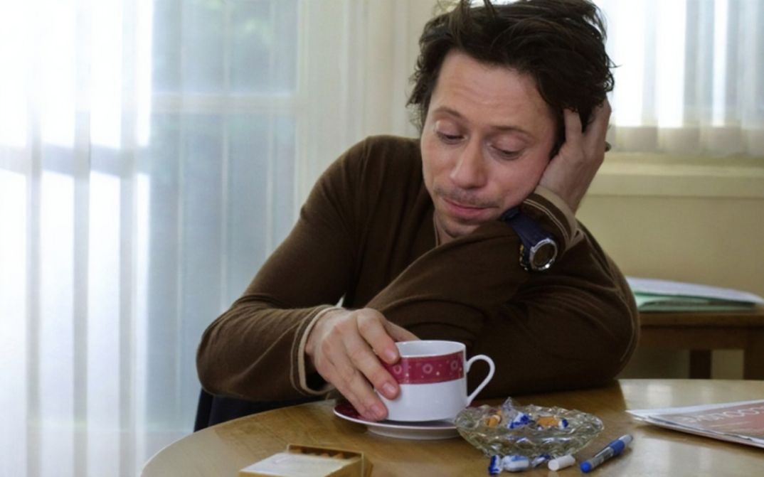 Filmstill aus ROIS ET REINE. Mathieu Amalric sitzt an einem Tisch, vor sich eine Kaffeetasse.